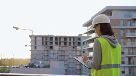 Woman engineer with a hard hat and safety vest is writing on a clipboard while inspecting a construction site at sunsetの写真素材