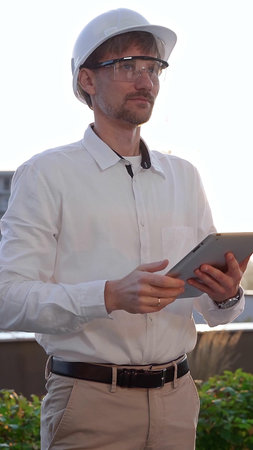 Male architectural engineer, wearing a white shirt and hard hat, is using a digital tablet while...