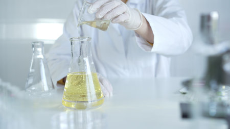 A scientist, wearing a lab coat and white protective gloves, is pouring a yellow oily liquid from one beaker to another in laboratory, close up. Medicine and science conceptの写真素材