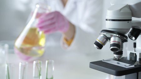 Microscope is near a scientist, wearing a lab coat and pink protective gloves, and holding beaker with a yellow oily liquid in laboratory setting, close up. Medicine and scienceの写真素材