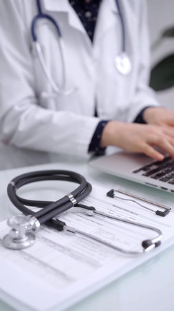 Stethoscope is lying on the glass table while doctor woman is working with laptop computer on the...