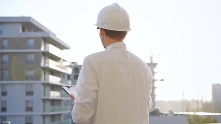 Man construction engineer wearing white shirt and hard hat is making notes on a clipboard while inspecting a building site at sunset, back view. Architecture and engineeringの写真素材