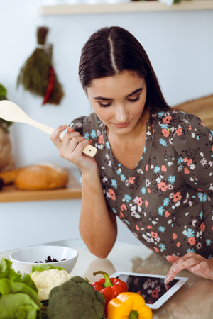 An attractive young dark-haired woman tastes a new recipe for a delicious salad mix while sitting at the table in the kitchen. Tablet pc is the best cookbookの写真素材