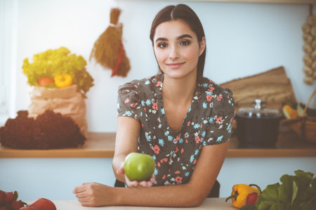 An attractive young dark-haired woman holding green apple and choosing the recipe for a delicious meal while smiling in sunny kitchen. Tablet pc is the best cookbookの写真素材