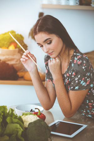 An attractive young dark-haired woman tastes a new recipe for a delicious salad mix while sitting at the table in sunny kitchenの写真素材