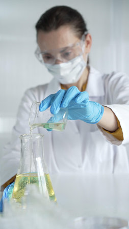 Woman scientist, wearing a lab coat, blue gloves and protective glasses, is pouring a yellow liquid...