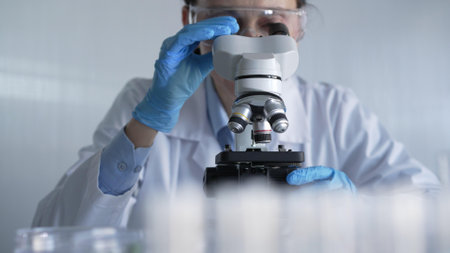 Woman scientist wearing lab coat, white protective gloves and glasses, is using microscope in modern laboratory setup, portrait view. Low key lighting photo. Science and Medicineの写真素材