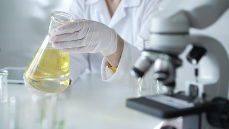 Microscope is near a scientist, wearing a lab coat and white protective gloves, and holding beaker with a yellow oily liquid in laboratory setting, close up. Medicine and science conceptの写真素材