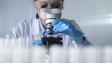 Woman scientist wearing lab coat, white protective gloves and glasses, is using microscope in modern laboratory setup, portrait view. Low key lighting photo. Science and Medicineの写真素材