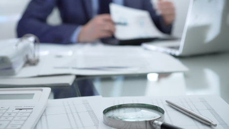 Calculator, pen, laptop and documents are lying on accountants desk. Businessman wearing blue suit is working with documents on the background. Business people, audit conceptの写真素材