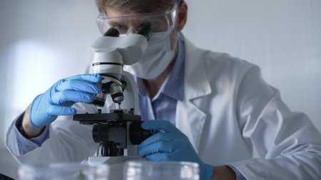 Male scientist researcher wearing blue gloves, protective glasses and medical mask, is using microscope in modern laboratory, portrait view. Low key lighting photo. Science and Medicineの写真素材