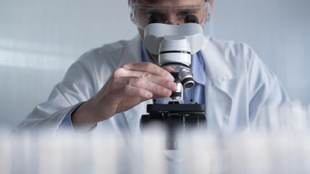 Male scientist wearing lab coat and protective glasses using microscope doing research in modern...