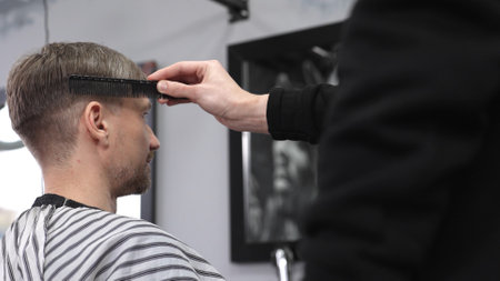 Barber carefully combing hair, client wearing protective cape, preparing for precision styling inside sleek contemporary barbershop settingの写真素材