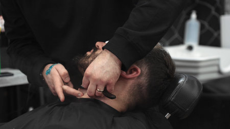 Professional barber carefully shaving clients beard with straight razor, applying gel and using hot towel for precise grooming in contemporary barbershop settingの写真素材