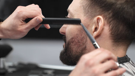 Skilled barber carefully trimming clients beard with precision scissors and sharp comb, working inside contemporary grooming salonの写真素材