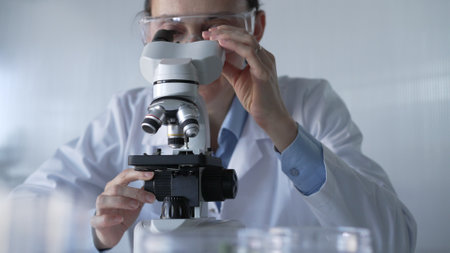 Woman scientist wearing lab coat and protective glasses is using microscope in modern laboratory setup. Low key lighting photo. Science and Medicineの写真素材
