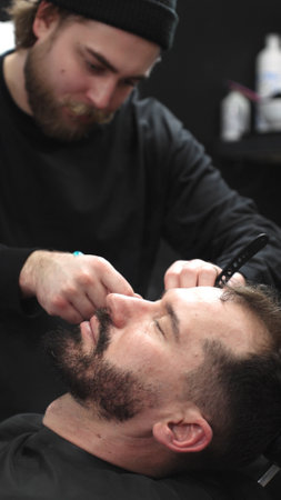 Professional barber carefully shaving clients beard with straight razor, applying gel and using hot towel for precise grooming in contemporary barbershop setting, vertical viewの写真素材