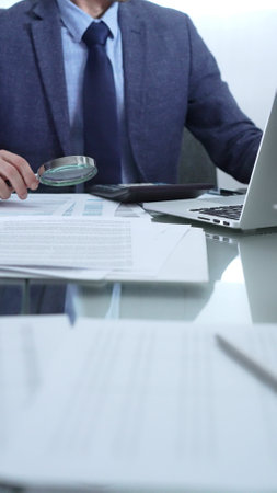 Businessman wearing blue suit is analyzing financial reports with magnifying glass and using calculator in a professional office setting. Low lighting, close-up vertical view. Business people, auditの写真素材