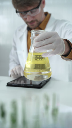 Man scientist wearing white protective gloves, and glasses is holding a yellow chemical solution inside an Erlenmeyer flask in a laboratory, vertical portraitの写真素材
