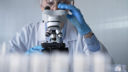 Woman scientist wearing lab coat, white protective gloves and glasses, is using microscope in modern laboratory setup, portrait view. Low key lighting photo. Science and Medicineの写真素材