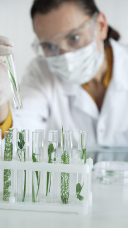 Woman scientist with white gloves, face mask and protective glasses, is holding a test tube with plants inside in laboratory setting, vertical view. Science and medicineの写真素材