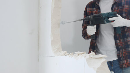 Close up of unknown male construction worker wearing red checkered shirt jeans and protective gloves, is demolishing white wall with rotary hammer drill, generating dust. Renovation conceptの写真素材