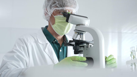 Medical scientist wearing protective mask, cap and green gloves using microscope doing scientific...