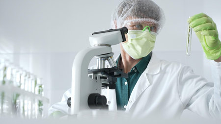Adult male scientist wearing green protective gear looking at plant samples in test tubes and using a microscope in modern laboratory for botanical research. Medicine, healthcare and science conceptの写真素材