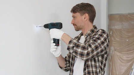 Middle aged man wearing checked shirt and white gloves, is drilling hole in a wall with cordless...