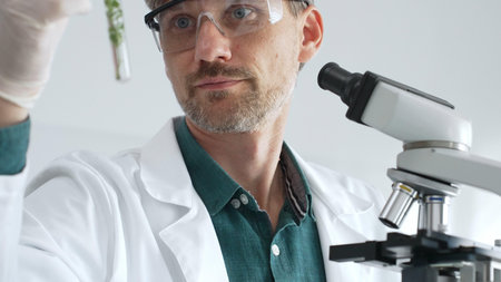 Adult male scientist in mask and gloves holding test tube with green plant sample, conducting...