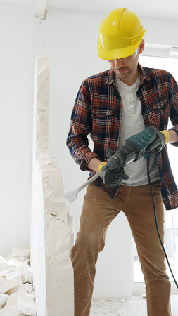 Male construction worker wearing red checkered shirt, yellow hard hat and protective gloves, is...