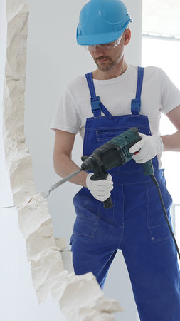 Man construction worker wearing blue overalls, hard hat and protective white gloves, is demolishing...