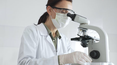 Female scientist examining microscope slide, wearing protective gear while conducting medical...