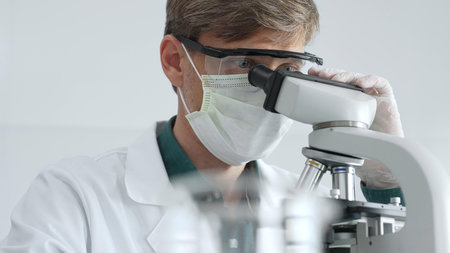 Masked adult scientist analyzing microscope slides, wearing green protective gear while conducting...