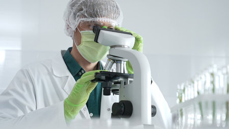 Medical scientist wearing protective mask, cap and green gloves using microscope doing scientific...