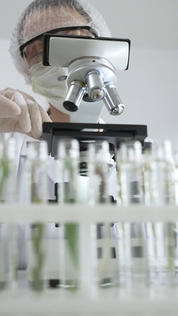 Scientist wearing green protective gear using microscope, analyzing test tubes in laboratory,...