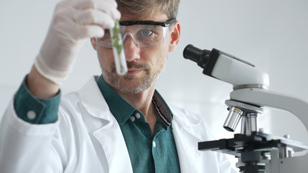 Adult male scientist in a lab coat and gloves examining a plant sample in a test tube, with a...