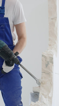 Unknown male construction worker wearing blue overalls and protective white gloves, is demolishing...