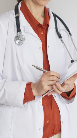 Close up of woman doctor wearing stethoscope and white coat over brick red blouse is standing and writing on a clipboard, representing medical expertise and patient care. Medicine conceptの写真素材