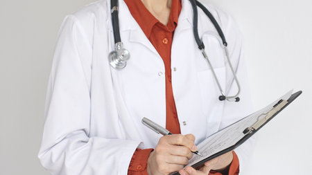 Unknown woman doctor wearing stethoscope and white coat over brick red blouse is standing and...