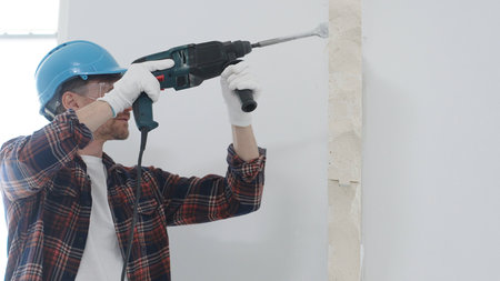 Male construction worker wearing red checkered shirt, blue protective helmet and gloves, is demolishing white wall with rotary hammer drill, generating dust, portrait view. Renovation conceptの写真素材