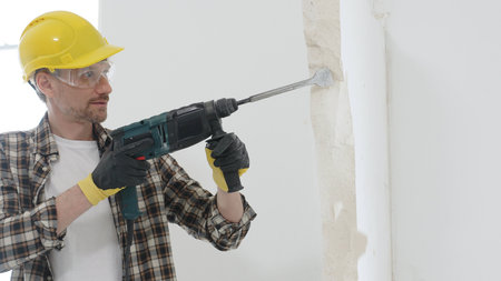 Portrait of a man construction worker wearing beige checkered shirt , yellow hard hat and protective gloves, is demolishing white wall with rotary hammer drill, generating dust. Renovation conceptの写真素材