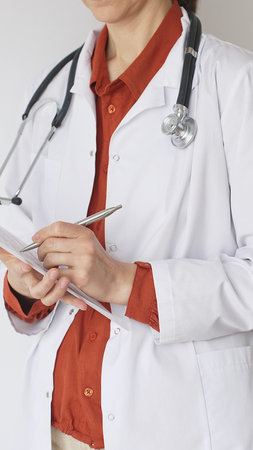 Close up of woman doctor wearing stethoscope and white coat over brick red blouse is standing and writing on a clipboard, representing medical expertise and patient care. Medicine conceptの写真素材