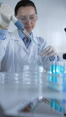 Female scientist wearing lab coat, white medical gloves and protective glasses is using micropipette...