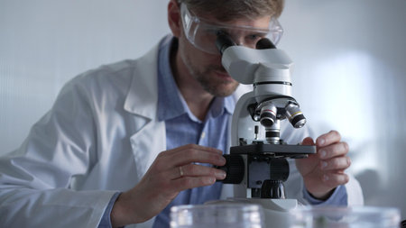 Male scientist wearing lab coat and protective glasses using microscope doing research in modern...