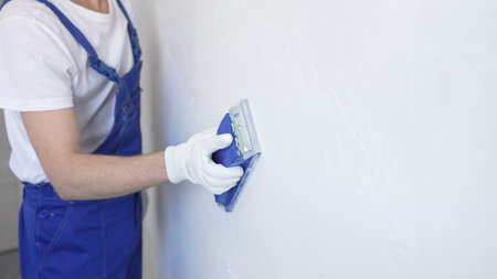 Unknown male construction worker wearing protective white gloves and blue construction coveralls, is sanding white wall with blue hand block, preparing smooth surface during interior home renovation projectの写真素材