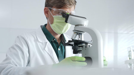 Adult male scientific professional analyzing sample through microscope, wearing green protective...