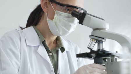 Female scientist examining microscope slide, wearing white protective gear while conducting medical...