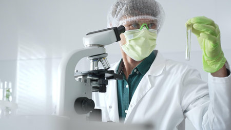 Adult male scientist wearing green protective gear looking at plant samples in test tubes and using...