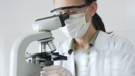 Female scientist examining microscope slide, wearing white protective gear while conducting medical...